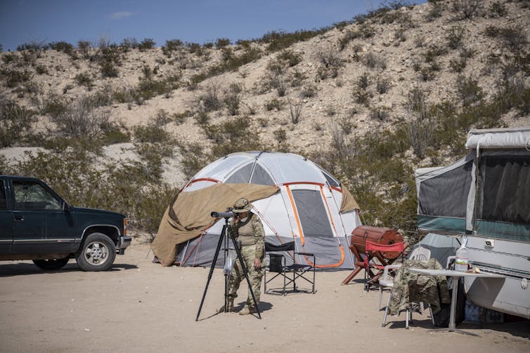 A man stands in front of a tent looking through a spotting scope.