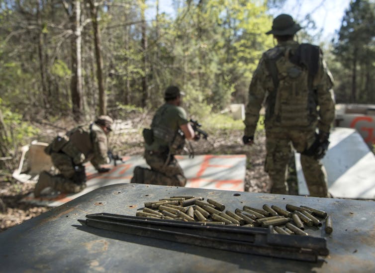 People in camouflage clothing hold weapons in a wooded area. Bullets are visible on a metal surface in the foreground.