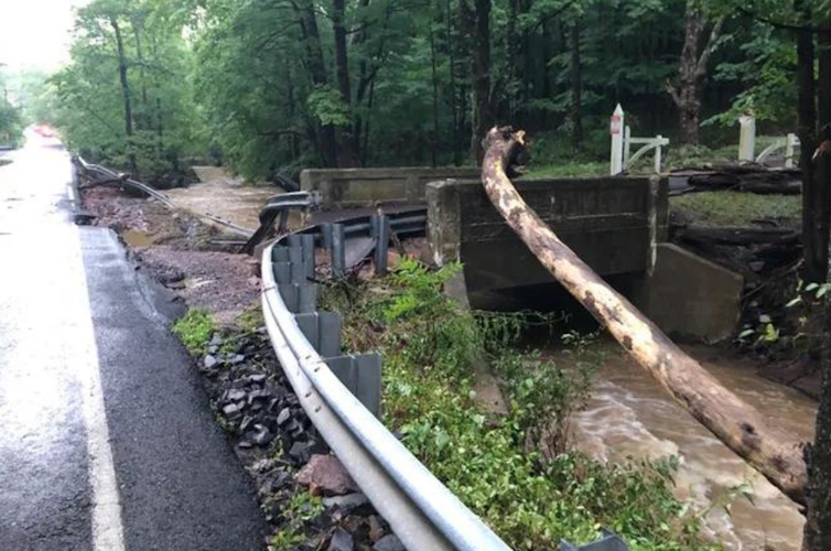 A small stream running beside a road, with toppled trees and damage to the road from flooding.