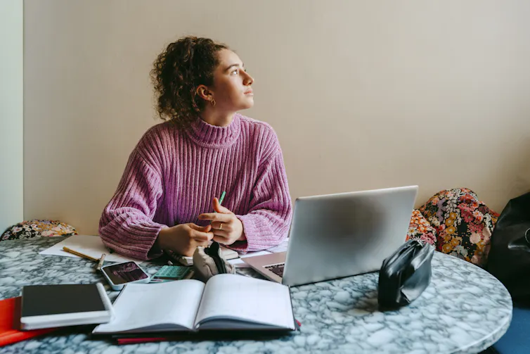 A young woman in a pink turtleneck sits in front of open books and an open laptop at an indoor table.