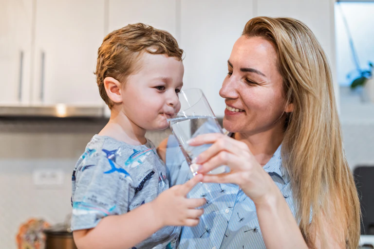 A toddler takes a sip of water as his mother holds a glass for him.