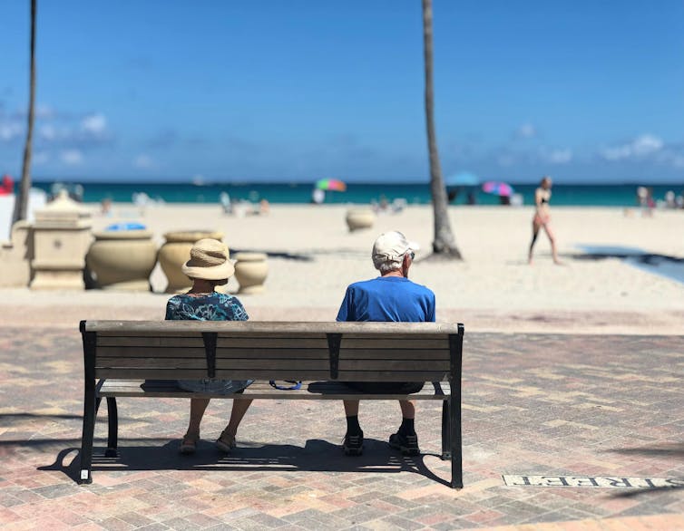 Two retirees sit on a bench by a beach.