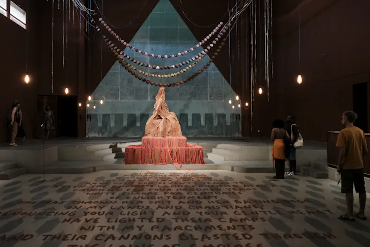 In a pool of light in a small amphitheatre with writing all along the floor leading up to it, orange and red cloth and rags cover a typical Victorian sculpture. Behind it, a triangle painted on a wall with flags hanging.