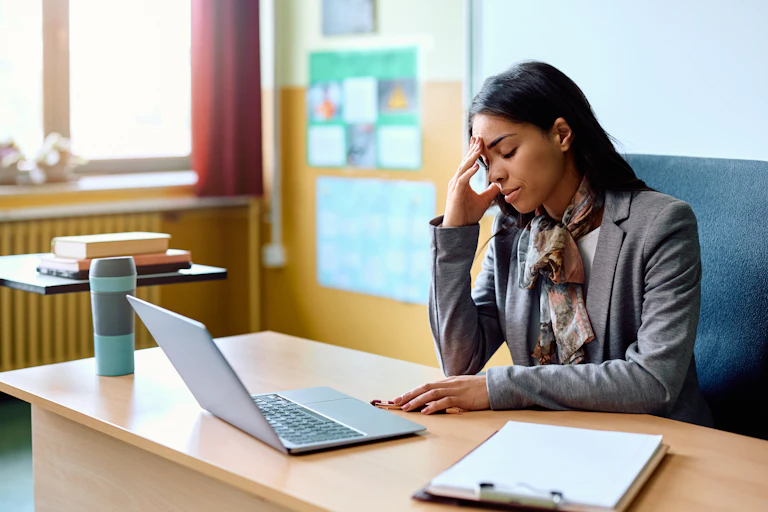 A teacher has her hand on her head as she sits at her desk with a laptop looking stressed.