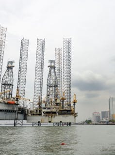 An oil rig with high spindly scaffolding towers in water with a dark cloud and a cityscape behind it.