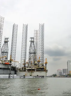 An oil rig with high spindly scaffolding towers in water with a dark cloud and a cityscape behind it.