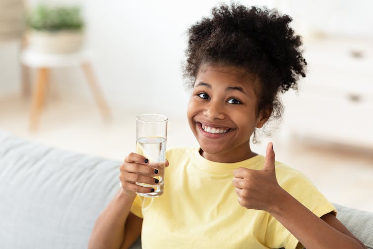 A teen in a yellow T-shirt holding a glass of water and giving the thumbs-up