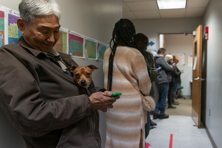A line of people in a building hallway, with the closest one looking at his phone and holding a small dog.