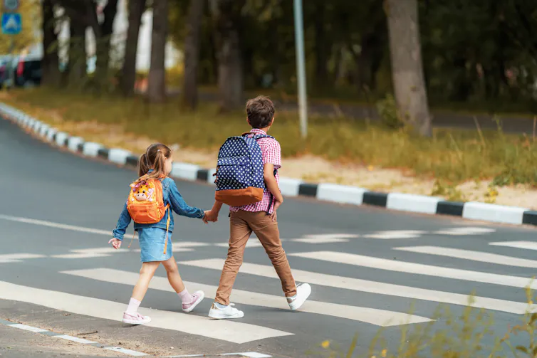 A boy and girl carrying backpacks hold hands as they walk along a crosswalk