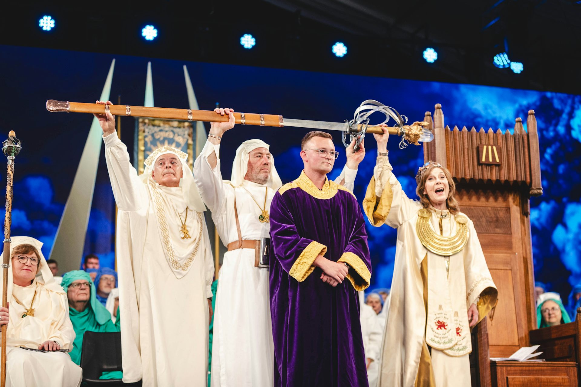 Three people in white robes hold a sword over the head of a man dressed in purple robes.