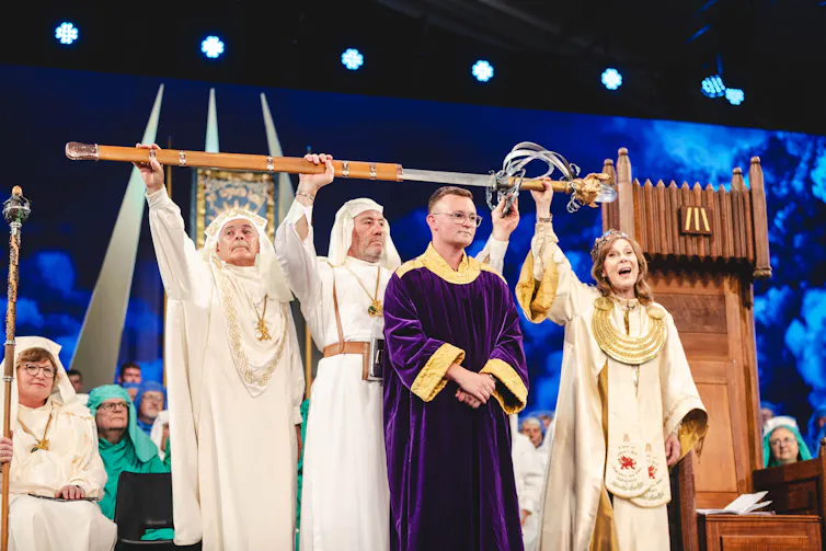 Three people in white robes hold a sword over the head of a man dressed in purple robes.