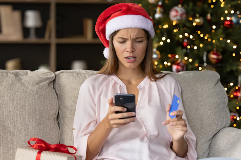 woman with long hair and christmas hat holding mobile and credit card, looking confused, christmas tree lit up in background