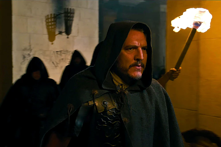 Pedro Pascal in hood walks through torch-lit corridor, with graffiti scrawled on the wall behind him