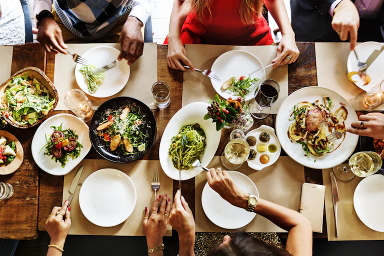 shot from above of people sat around table, hands helping themselves to food, white plates and cutlery