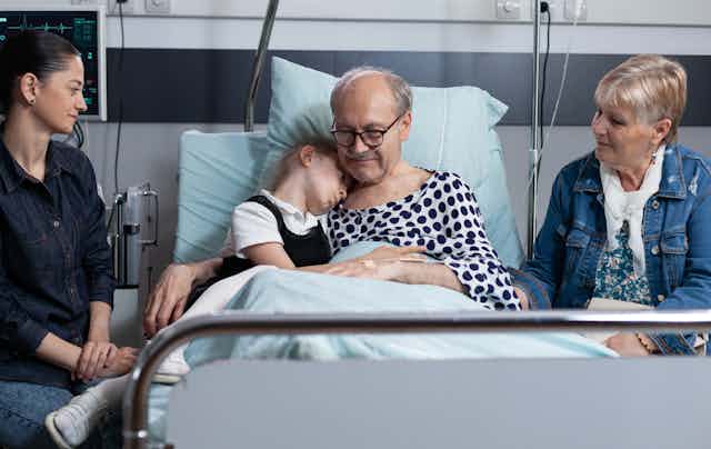 A family gathers around an older man in a hospital bed, with a young girl on the bed with him to give a hug