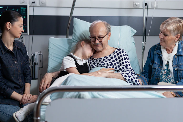 A family gathers around an older man in a hospital bed, with a young girl on the bed with him to give a hug