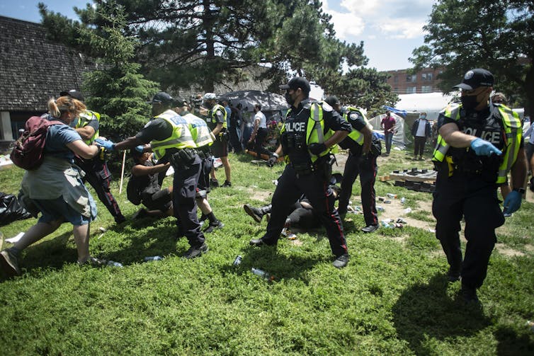 Police forcibly remove homeless encampment supporters on a sunny summer day.