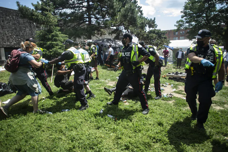 Police forcibly remove homeless encampment supporters on a sunny summer day.