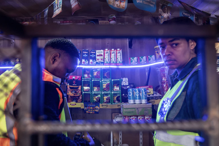 Two men in reflective vests inside a small ship where a few products are on shelves, seen through an opening in a wire fence