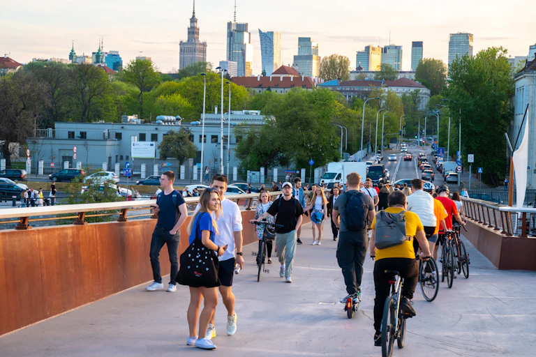 A group of people, some on bicycles, cross a bridge, with skyscrapers in the background.