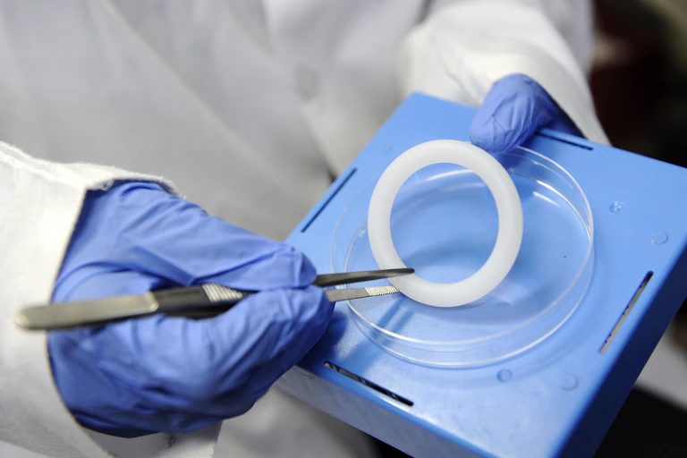 Hands wearing blue medical gloves holding a container and tweezers which grip a white ring-shaped object
