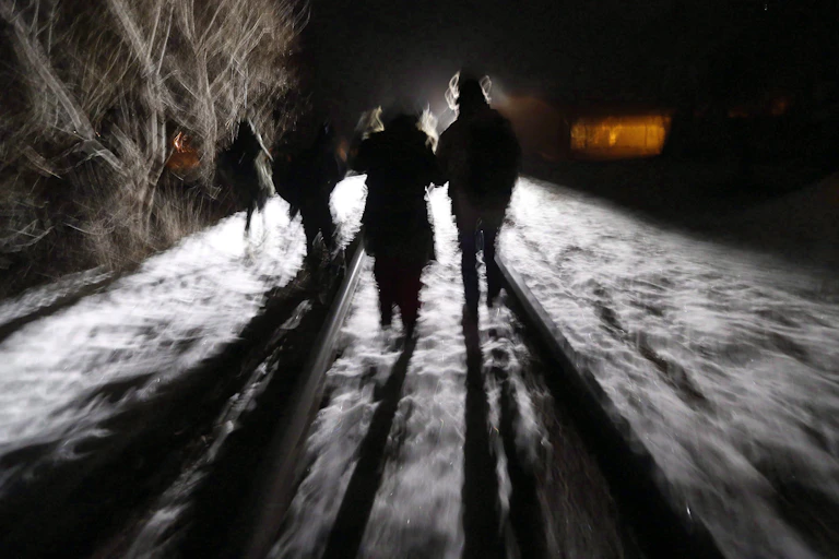 Migrants in silhouette are seen walking on a snowy night along a train tracks.