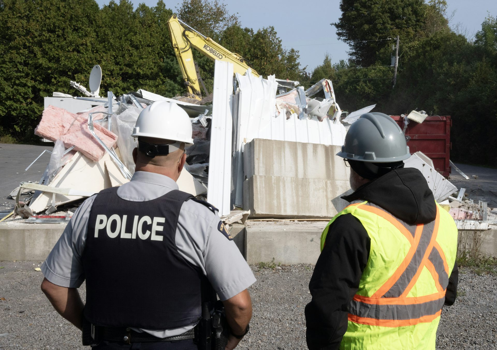 A police officer and a person in a yellow safety vest and a hard helmet watch the demolition of a building.