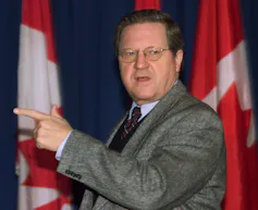 A dark-haired man in glasses points ahead of him with flags in a row behind him.