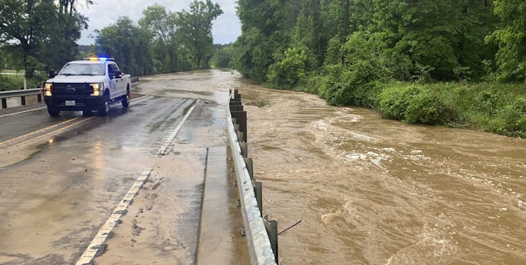 A truck is parked at the edge of a road to alert drivers of an area where muddy water is washing over the bridge beyond.