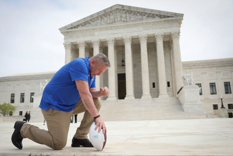 Man in blue polo shirt kneels on a stairwell in front of a large white building with columns.