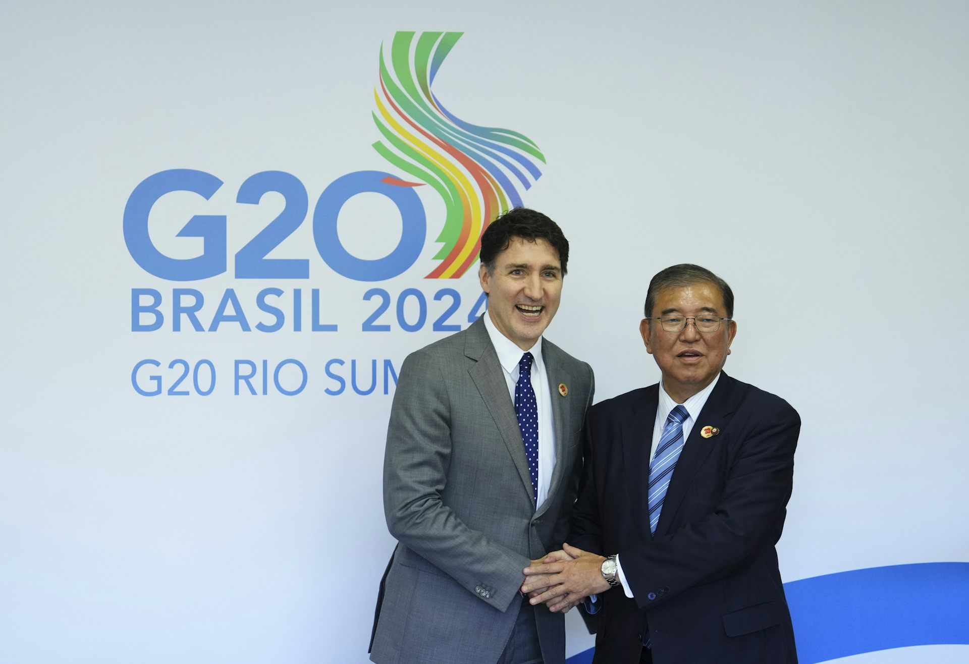 A dark-haired white man smiles as he shakes hands with a dark-haired Asian man. Behind them a banner reads G20 Brasil 2024.