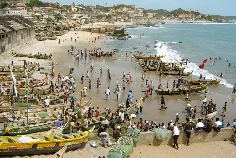 Fishing boats in front of Cape Coast Castle, Ghana, with homes and buildings along the coast.