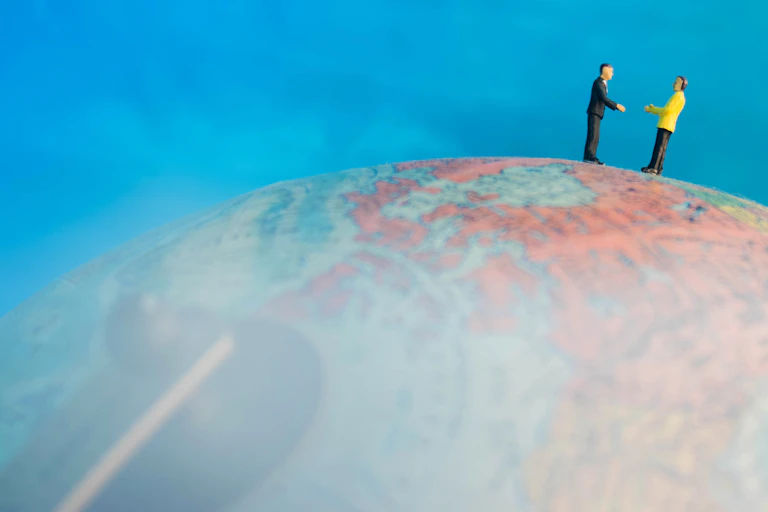 two figurines standing on globe, holding out hands, blue sky