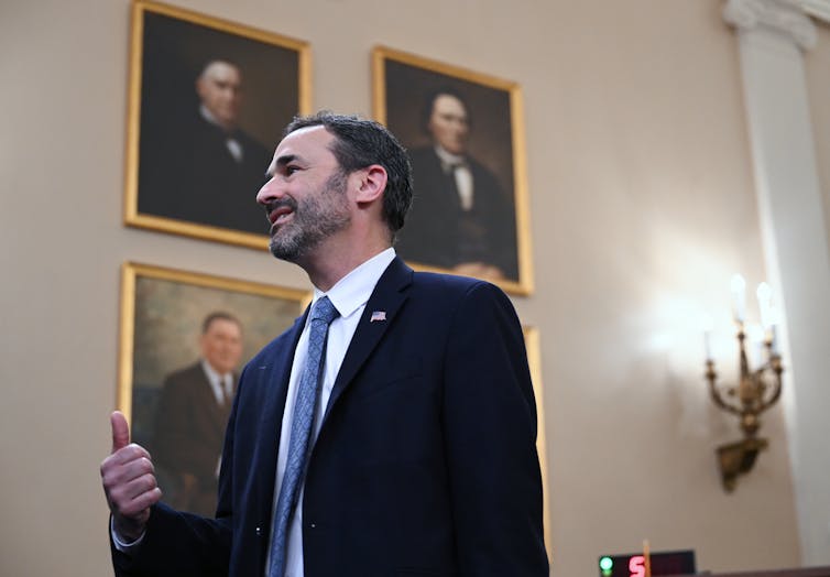 A bearded man wearing a blue blazer with an American flag pin and a blue neck tie and dress shirt stands in front of a wall of portraits while motioning with his hand during testimony before Congress.