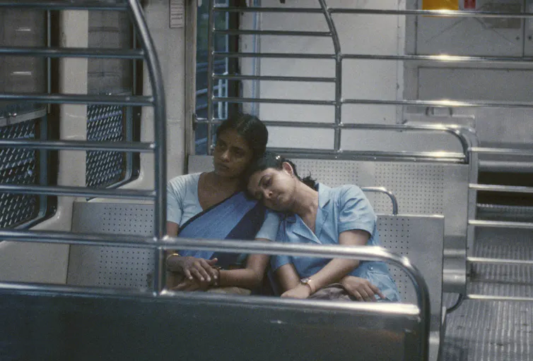 Two indian women in blue outfits sit on a train. One rests her head on the other's shoulder.
