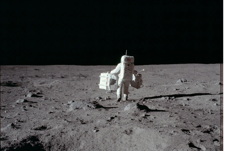 An astronaut bounds across the surface of the Moon while carrying bulky equipment.