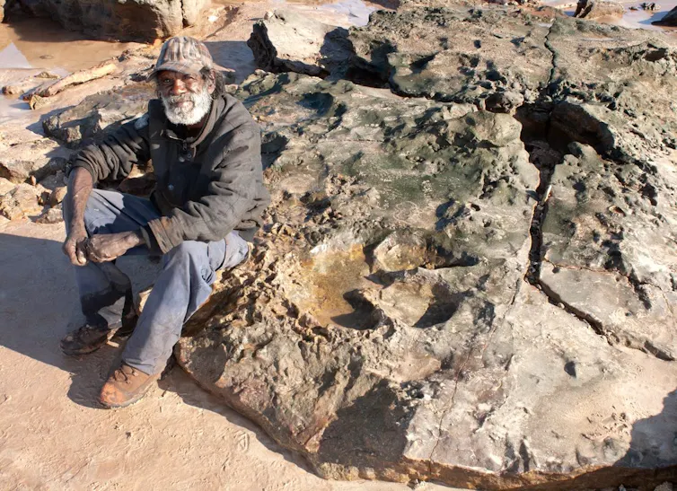 Photo of an Aboriginal man sitting on a large rock that carries the impression of a huge three-toed foot.