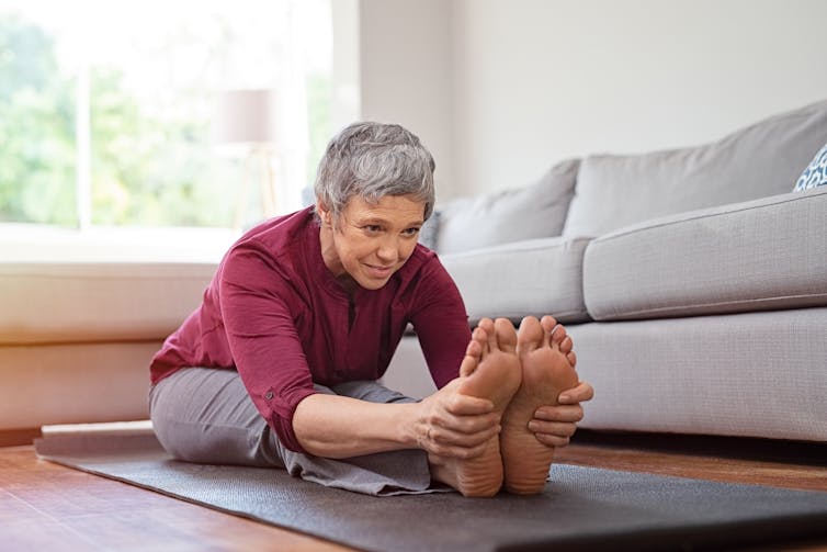 A senior woman stretching on a yoga mat in a living room.