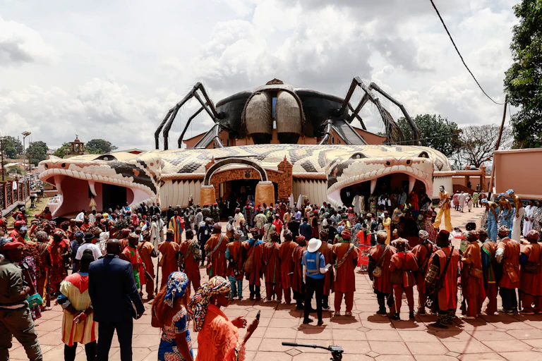 A crowd of people stands outside a curious building, with an entrance and exit on either side through a giant snake's head with fangs that wraps around the building with a huge hairy spider perched on the roof.