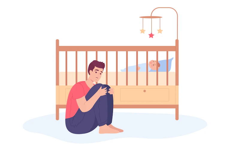 A father sits on the ground next to his infant's bed, looking worried.