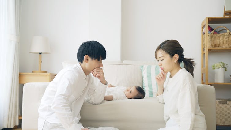 A couple has a serious discussion while their baby lays on the couch behind them.