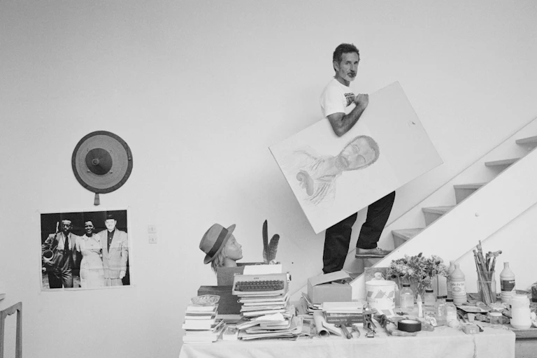A black and white image of a man with a beard carrying a portrait of himself under his arm up a flight of white stairs, looking at camera.