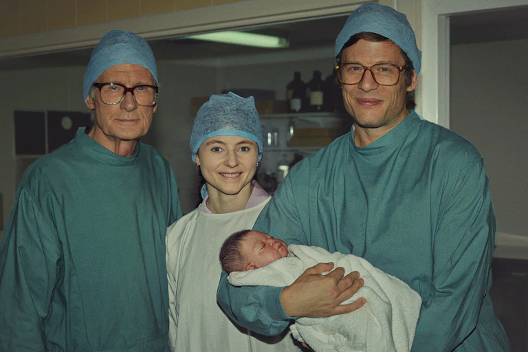 Bill Nighy, Thomasin McKenzie and James Norton dressed in scrubs holding a baby.