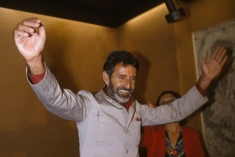 A smiling man with a beard holds both arms up in the air, festive.