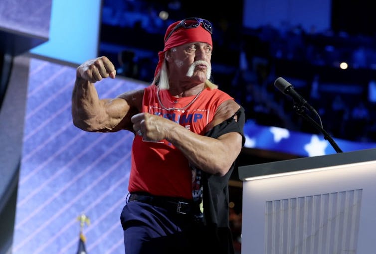 Older, muscled man with a handlebar mustache wearing a red banadana and red, sleeveless T-shirt flexes at a podium.