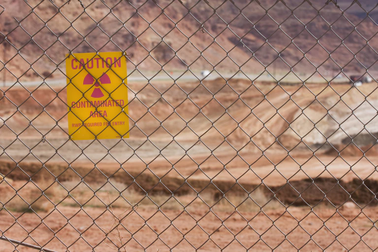A big sandy area seen through a wire fence. Hanging on the fence is a notice - caution, contaminated area