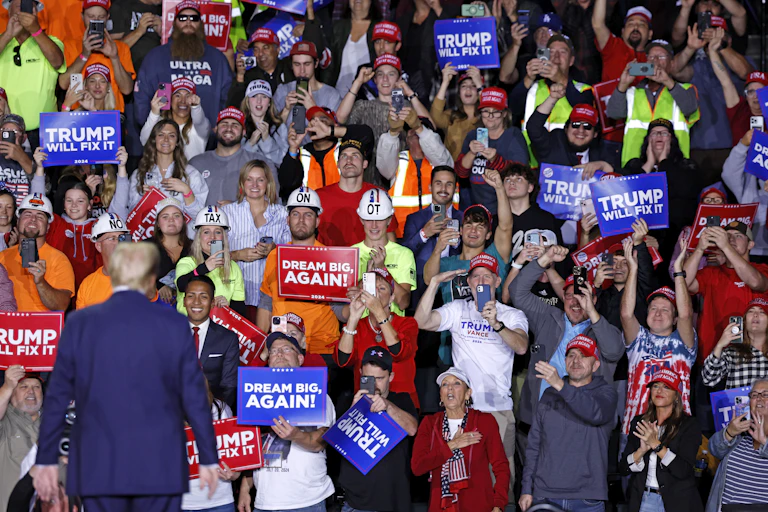 A man wearing a navy blue suit is seen from behind looking at a large crowd of people who hold red and blue signs that say 'Trump will fix it' and 'Dream big, again!'