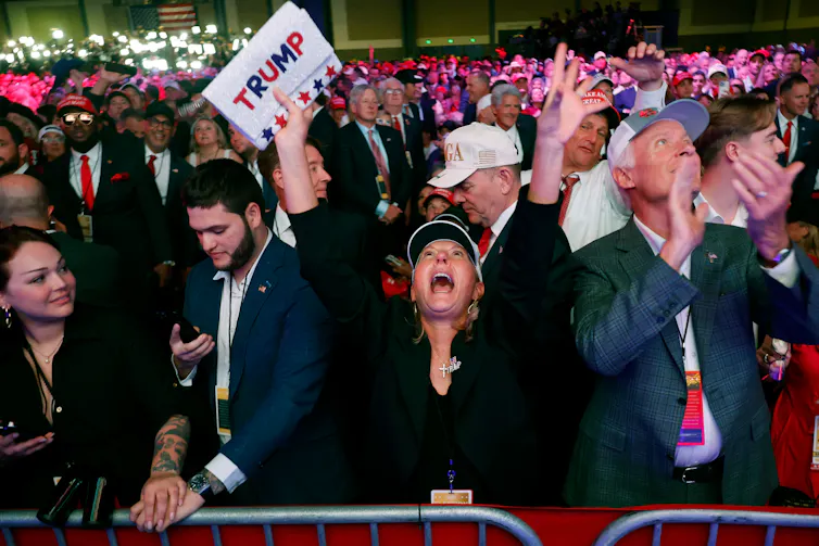 People stand in a large crowd and a woman in the center smiles broadly and has her arms in the air. She holds a white sign that says 'Trump'