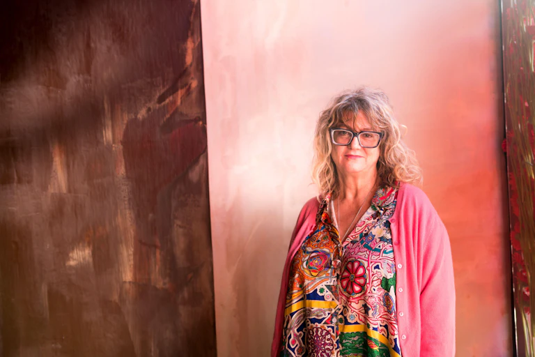 An woman artist in a brightly coloured dress and pink cardigan against a background of chocolate-painted walls.