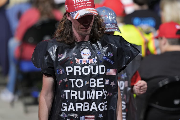 A man wears a red hat and a black trash bag that says in white writing, 'Proud to be Trump's garbage.'